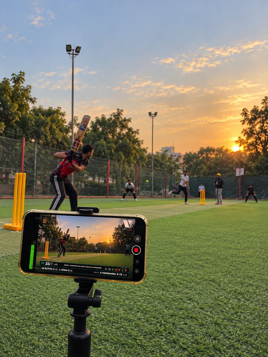 A batsman mid-shot at a turf ground, captured live on a phone in the foreground